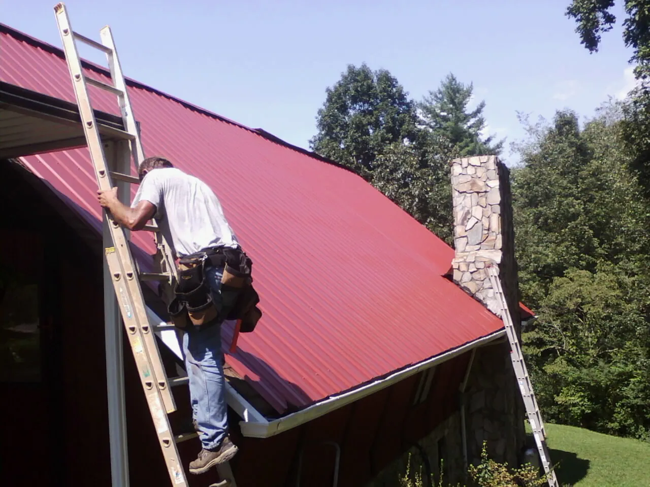 Roofer on a ladder beside a red metal roof and stone chimney — Soffit & Fascia Repair by Roofing Brothers in Mc Caysville